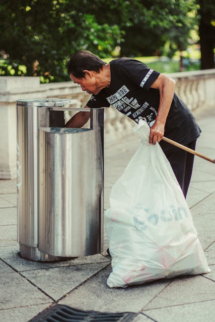 A middle-aged man with dark hair wearing a black T-shirt with white text and dark trousers is seen disposing of a large white plastic bag filled with rubbish into a stainless steel litter bin on a paved urban sidewalk. The man is leaning forward, holding a wooden stick or litter picker in his left hand and gripping the bag with his right hand as he empties it into the bin’s open slot. The stainless steel bin has a polished finish, with vertical panels and a rounded top section. In the background, a low stone wall runs parallel to the sidewalk, and lush green foliage and trees provide shade, suggesting an outdoor park or walkway area. The scene is illuminated by natural daylight, ensuring clear visibility of textures, colors, and the environmental context. This image relates to private waste collection and on-site rubbish disposal, emphasizing the importance of proper waste management and alternative rubbish removal methods provided by waste clearance services like wasteclearancequeenspark.co.uk.