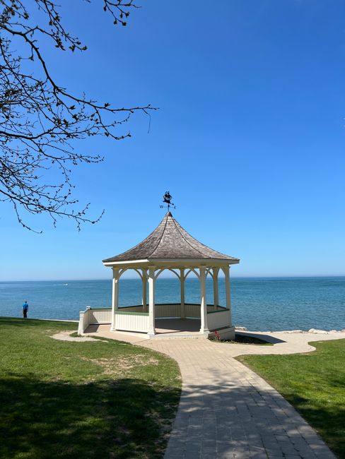 A white, open-sided gazebo with a shingled, conical roof and decorative weather vane on top is situated on a grassy area near the shoreline, overlooking the sea under a clear blue sky. The gazebo is positioned slightly to the right of the center of the image, with a paved pathway leading up to it from the foreground. To the left, a bare tree branch extends into the frame, partially shading the area. In the background, the calm ocean stretches out to the horizon, with a solitary person dressed in blue standing near the water's edge on the left side. The scene appears peaceful and quiet, with natural light highlighting the textures of the grass, pathway, and wooden structure, giving a sense of a well-maintained outdoor space that could be associated with leisure or public seaside settings while subtly aligning with outdoor waste and rubbish removal contexts through the clean, orderly environment.