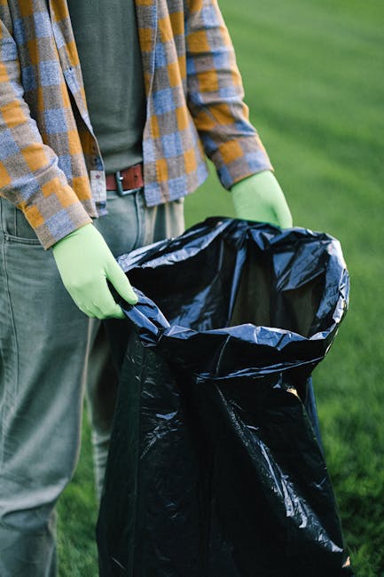 A middle-aged man with dark hair wearing a black T-shirt with white text and dark trousers is seen disposing of a large white plastic bag filled with rubbish into a stainless steel litter bin on a paved urban sidewalk. The man is leaning forward, holding a wooden stick or litter picker in his left hand and gripping the bag with his right hand as he empties it into the bin’s open slot. The stainless steel bin has a polished finish, with vertical panels and a rounded top section. In the background, a low stone wall runs parallel to the sidewalk, and lush green foliage and trees provide shade, suggesting an outdoor park or walkway area. The scene is illuminated by natural daylight, ensuring clear visibility of textures, colors, and the environmental context. This image relates to private waste collection and on-site rubbish disposal, emphasizing the importance of proper waste management and alternative rubbish removal methods provided by waste clearance services like wasteclearancequeenspark.co.uk.