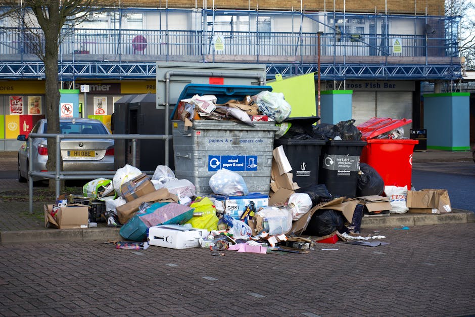 A large collection of mixed waste and rubbish is piled up on a paved surface in front of a row of refuse bins, with some cardboard boxes, paper bags, plastic bags, and loose packaging materials visible. The rubbish includes various discarded packaging, soft plastics, and paper products, some spilling onto the surrounding area. Behind the waste, a grey metal recycling container marked for mixed paper and cardboard is partially open, and a red bin is positioned nearby. To the left, a small green railing separates the rubbish from a parked silver car with a UK license plate. In the background, a building with large windows, blue scaffolding, and shopfronts, including a fish bar, can be seen, situating the scene in an urban environment. The area is illuminated by natural daylight, with some shadowing from trees and surrounding structures, reflecting a typical waste collection or disposal scene suitable for a UK rubbish removal service context.