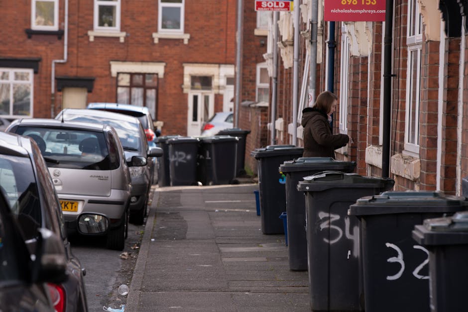 The image shows a row of dark grey wheelie bins placed along the pavement in front of a brick residential building. Several bins have white painted numbers on their sides, including the numbers '32' and '34.' A woman dressed in a brown coat is standing next to the bins, appearing to be disposing of rubbish. The building has white-framed windows and a partially visible red 'SOLD' sign hanging from the wall. Parked cars, including a silver hatchback, line the street on the left side, with some vehicles facing towards the background and others partially visible in the foreground. The street and pavement appear clean, and the environment suggests an urban residential area where residents manage waste collection privately, possibly as an alternative to council-led rubbish disposal. The scene is illuminated by natural daylight, with a clear atmosphere and no visible waste outside of the designated bins. The setting aligns with typical private rubbish collection areas in Queens Park, supported by local waste clearance services such as those offered by wasteclearancequeenspark.co.uk.